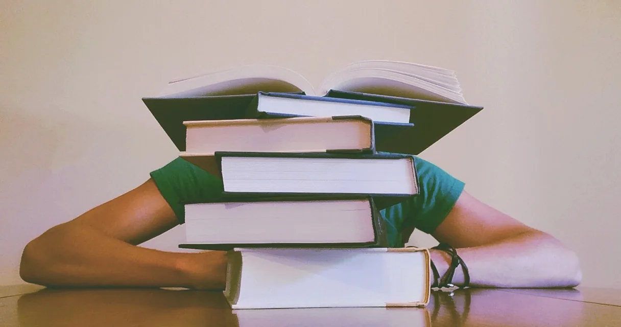 Pilha de livros sobre uma mesa, com uma pessoa debruçada atrás deles