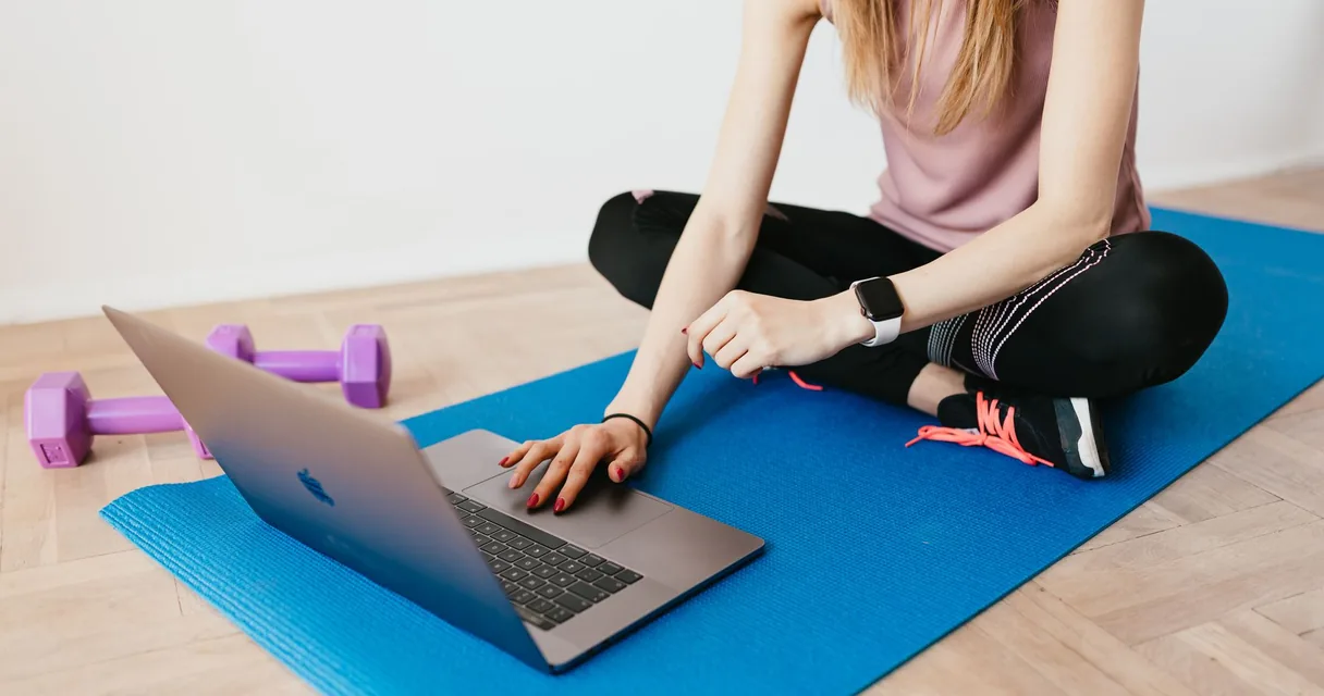 Mulher sentada no tapete de treino em casa usando notebook, com halteres ao lado