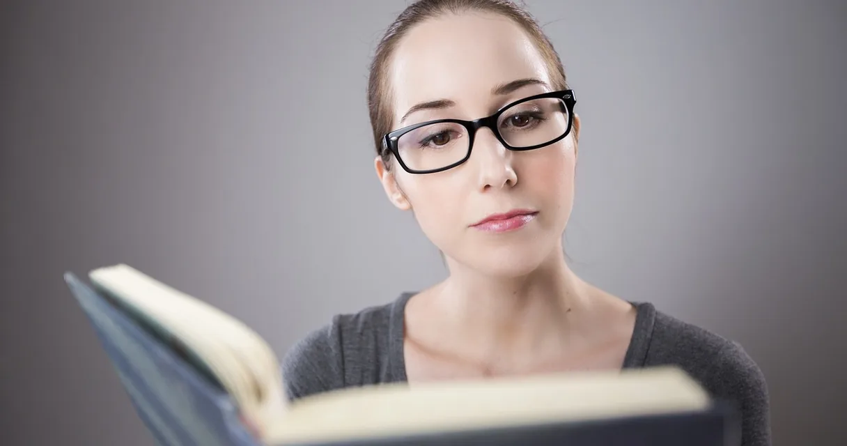 Mulher de óculos lendo um livro aberto diante de um fundo cinza