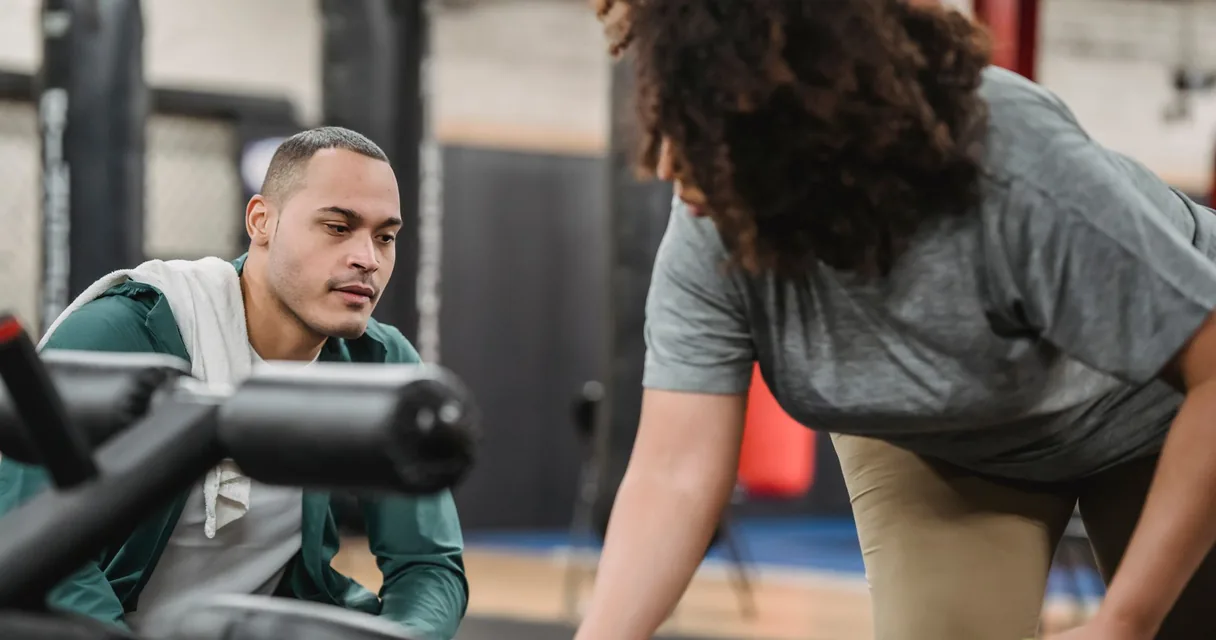 Personal trainer agachado orientando aluna durante exercício na academia